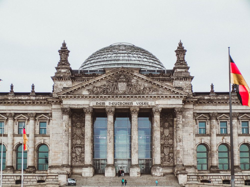 Close-up of the Reichstag building in Berlin, showing the detailed classical façade, columns, and the inscription "DEM DEUTSCHEN VOLKE" with a German flag on the right.